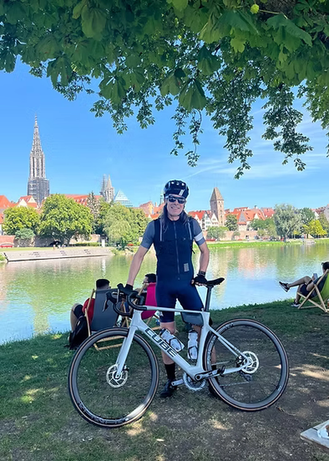 Der Laufcoach - Leistungscoaching und Analyse für Läufer - Ein Radfahrer steht mit einem weißen Cube-Rennrad an einem grasbewachsenen Ufer mit Blick auf einen Fluss; im Hintergrund sind das historische Ulmer Münster und das Stadtbild zu sehen.