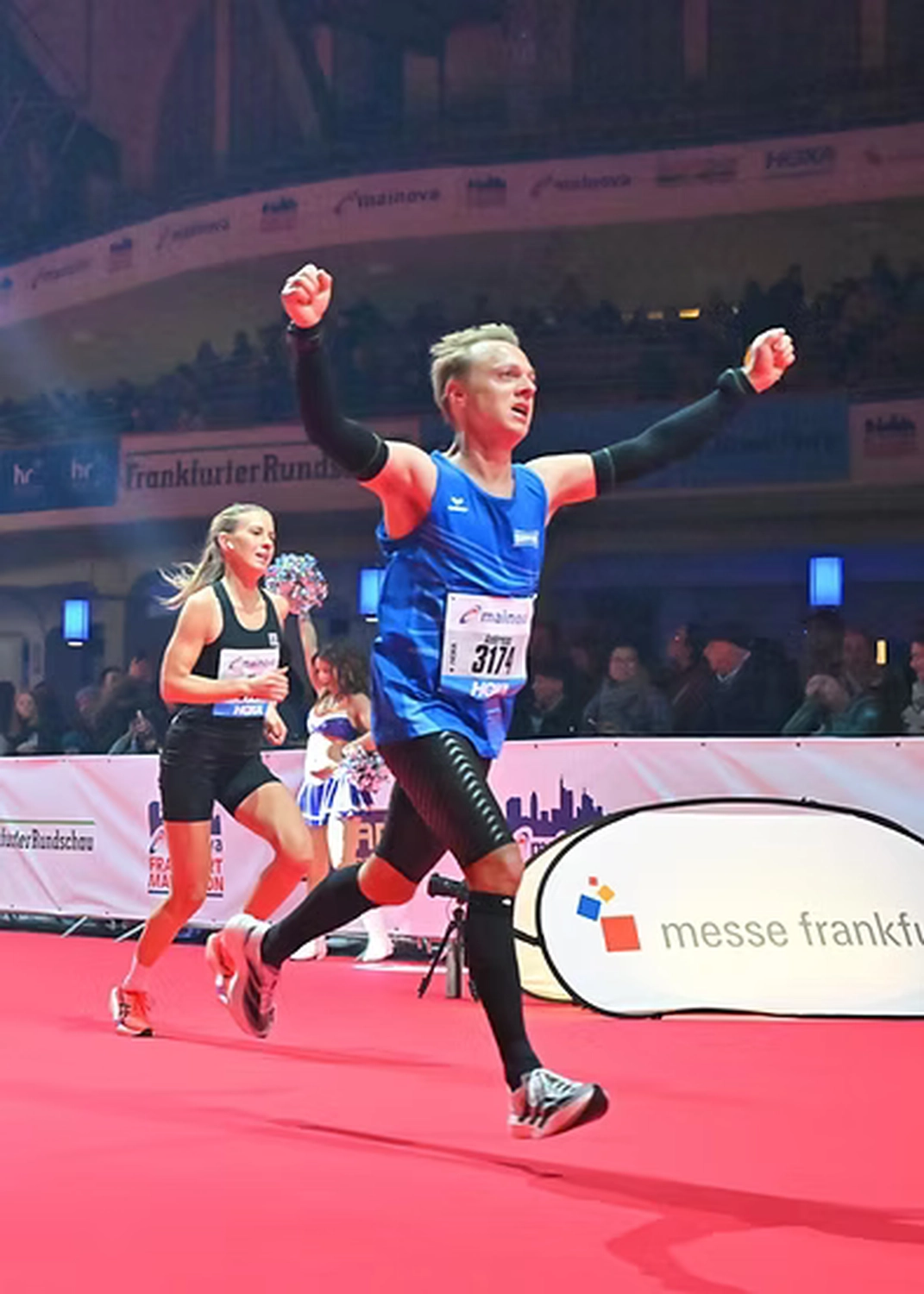Der Laufcoach - Leistungscoaching und Analyse für Läufer - Ein männlicher Läufer in einem blauen Tanktop und schwarzer Kompressionskleidung jubelt mit erhobenen Armen, als er auf einem roten Teppich die Ziellinie überquert; im Hintergrund sind eine Läuferin und eine Menschenmenge zu sehen.