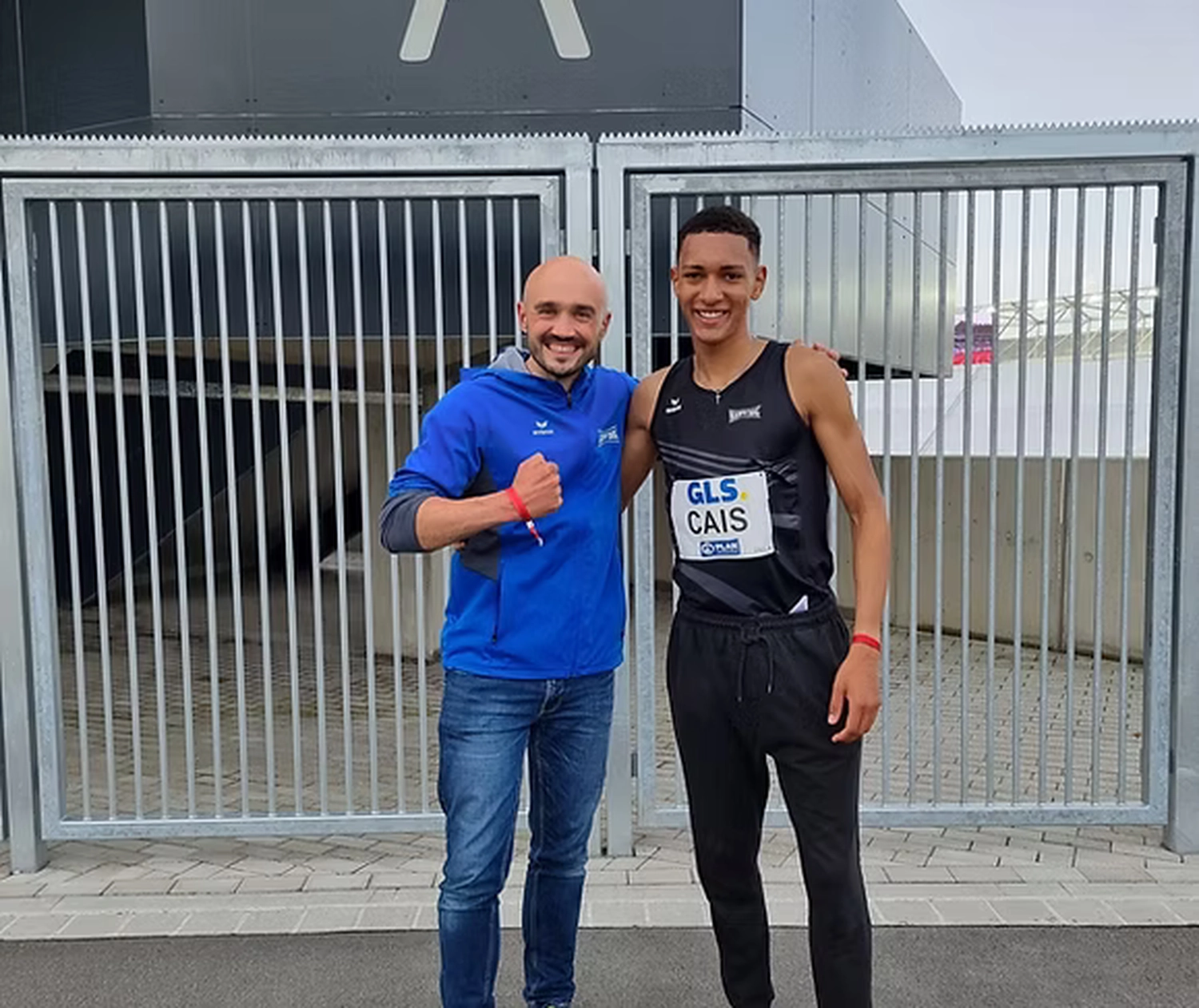 Der Laufcoach - Leistungscoaching und Analyse für Läufer - Zwei Männer stehen vor einem Metalltor vor einem modernen Gebäude; der Mann links trägt eine blaue Jacke, der Mann rechts einen schwarzen Trainingsanzug.