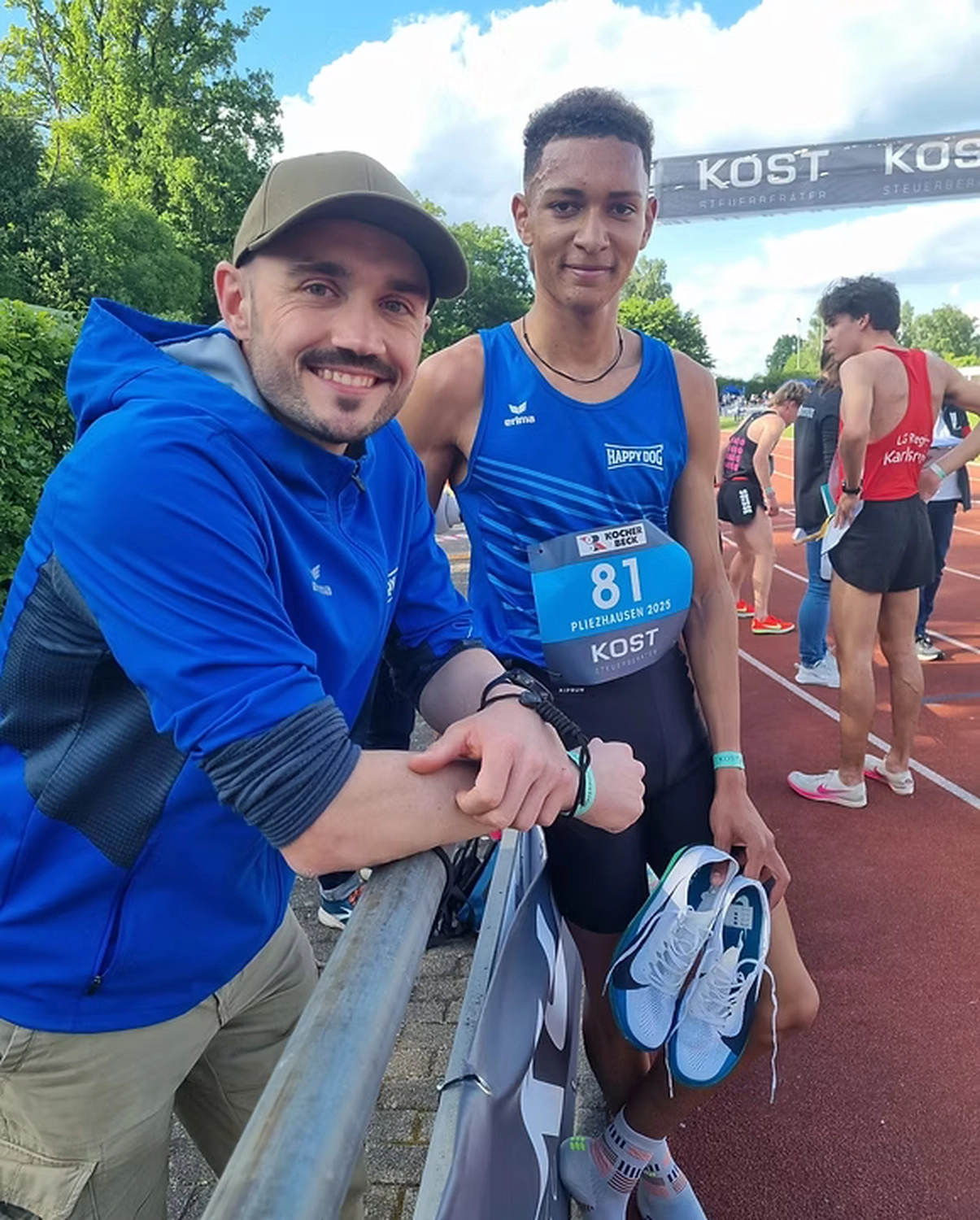 Der Laufcoach - Leistungscoaching und Analyse für Läufer - Ein lächelnder Mann in einem blauen Kapuzenpulli und einer olivgrünen Baseballkappe lehnt an einem Metallgeländer neben einem jungen Leichtathleten in einem blauen Tanktop, der seine Spikes hält.