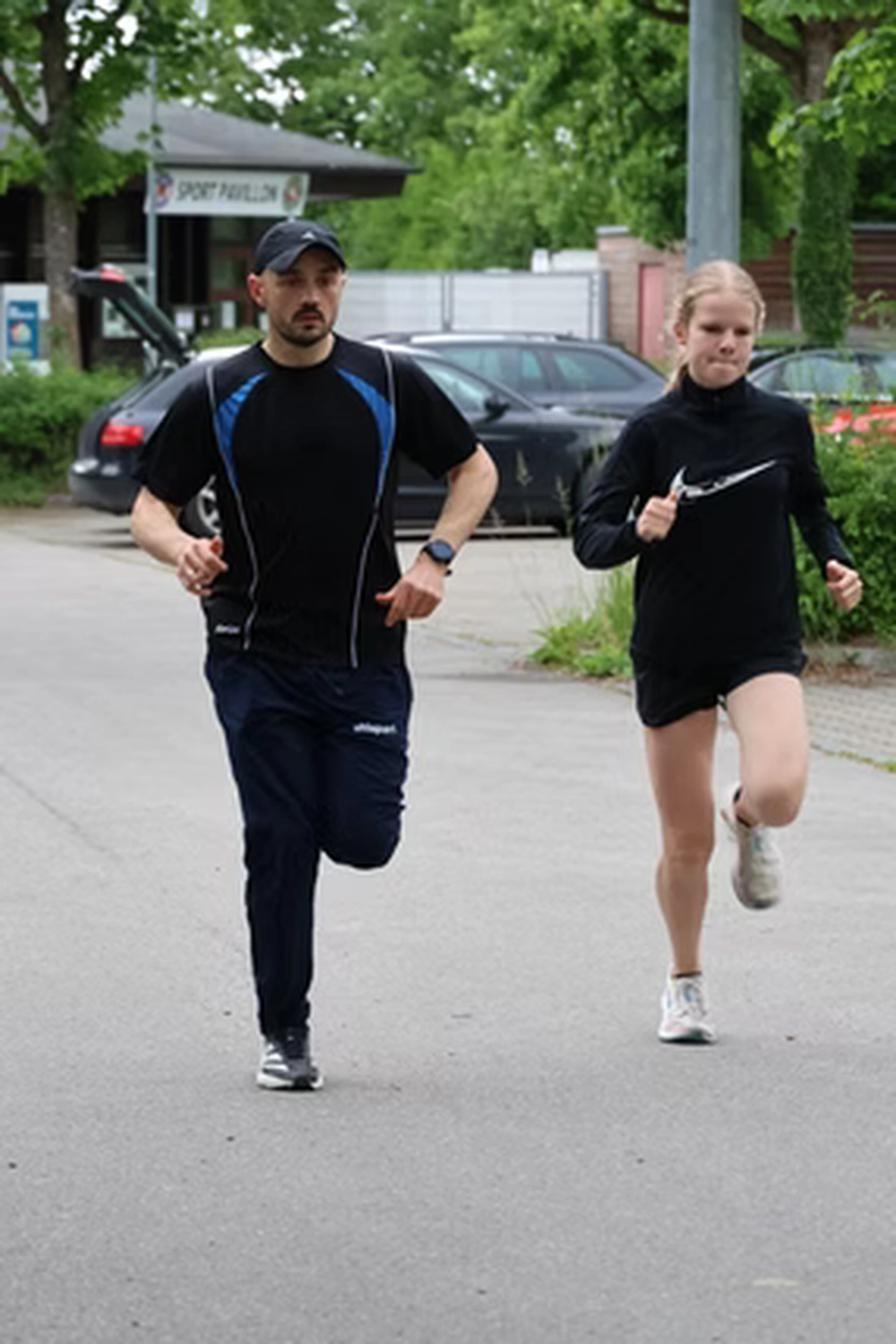Der Laufcoach - Leistungscoaching und Analyse für Läufer - Ein Mann und eine Frau in Sportkleidung rennen gemeinsam auf einer asphaltierten Straße; im Hintergrund sind Autos und Bäume zu sehen.