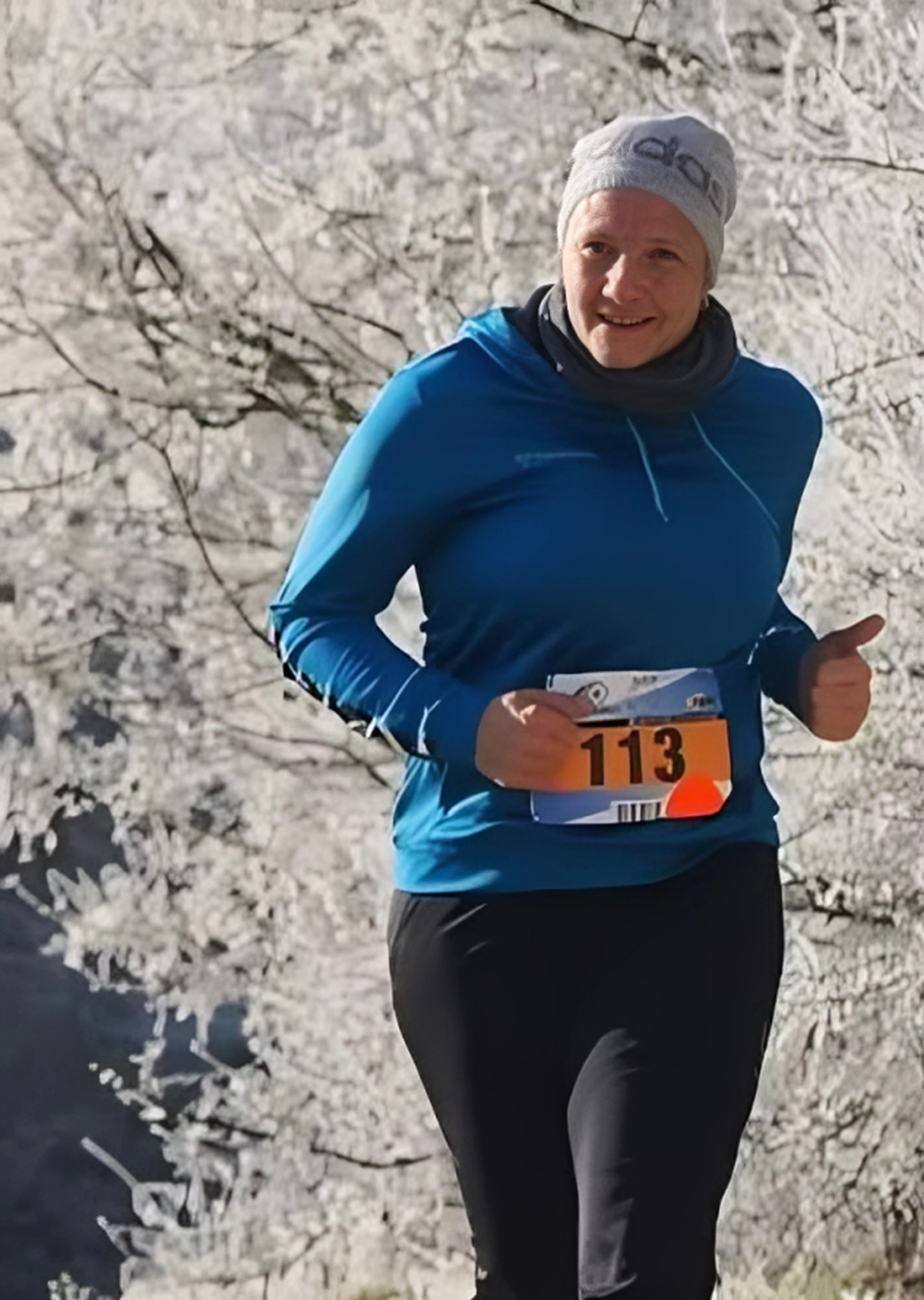 Der Laufcoach - Leistungscoaching und Analyse für Läufer - Eine Frau in blauem Kapuzenpullover, schwarzen Leggings und grauer Adidas-Mütze lächelt, während sie in einer verschneiten oder frostbedeckten Landschaft an einem Lauf teilnimmt. Sie trägt eine Startnummer mit der Nummer 113 an der Brust.