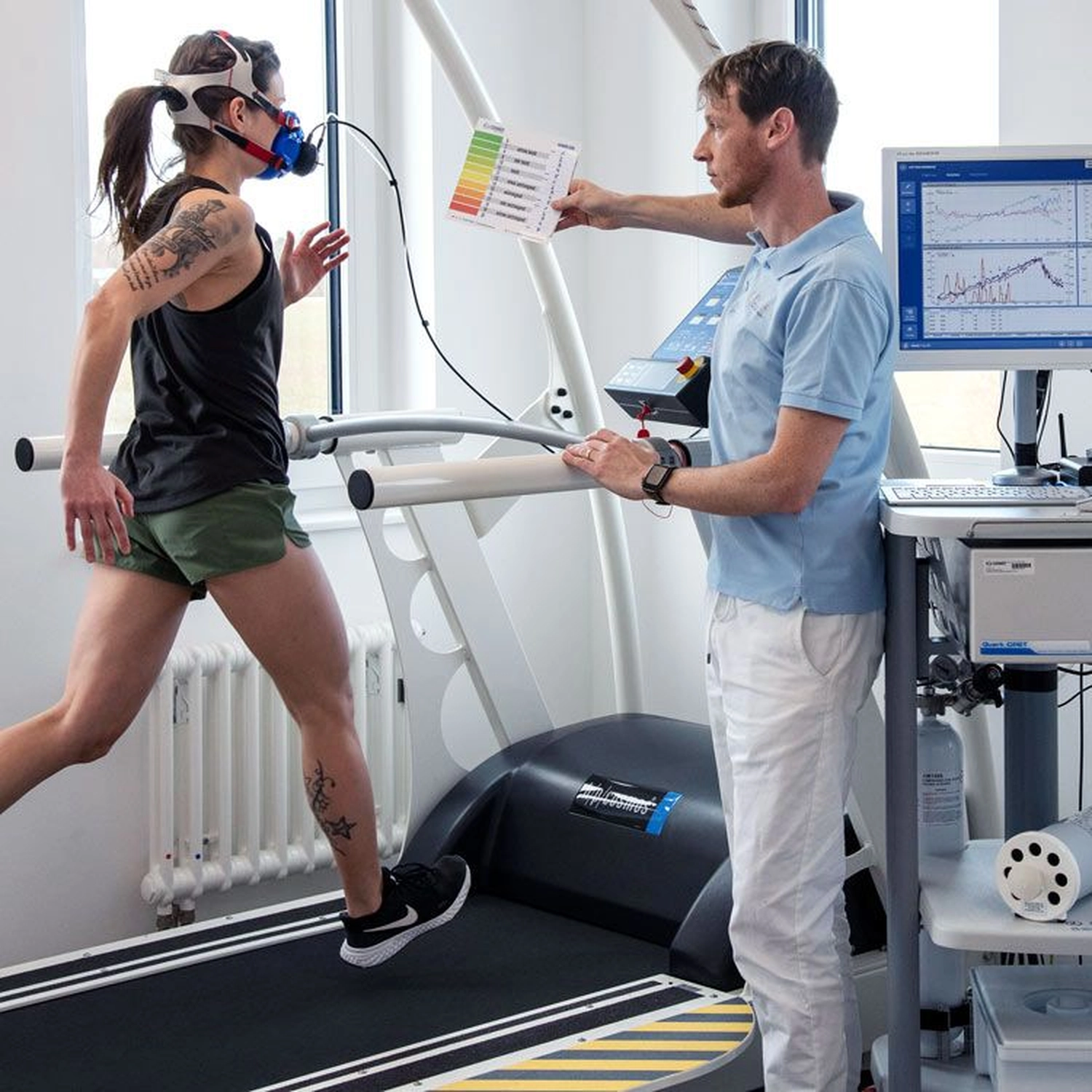 Der Laufcoach - Leistungscoaching und Analyse für Läufer - Eine Frau mit Atemschutzmaske läuft auf einem Laufband, während ein Techniker in einer Klinik ihre Vitalwerte auf einem Computerbildschirm überwacht.
