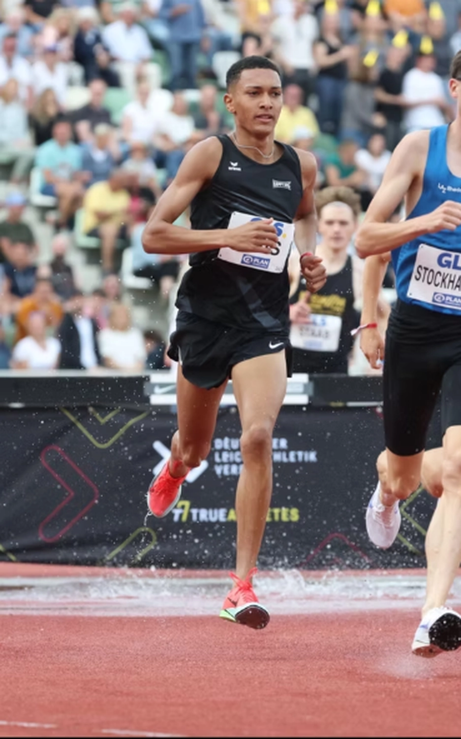 Der Laufcoach - Leistungscoaching und Analyse für Läufer - Ein männlicher Leichtathlet in schwarzem Tanktop und Shorts läuft während eines Wettkampfs auf einer roten Laufbahn durch Wasser.
