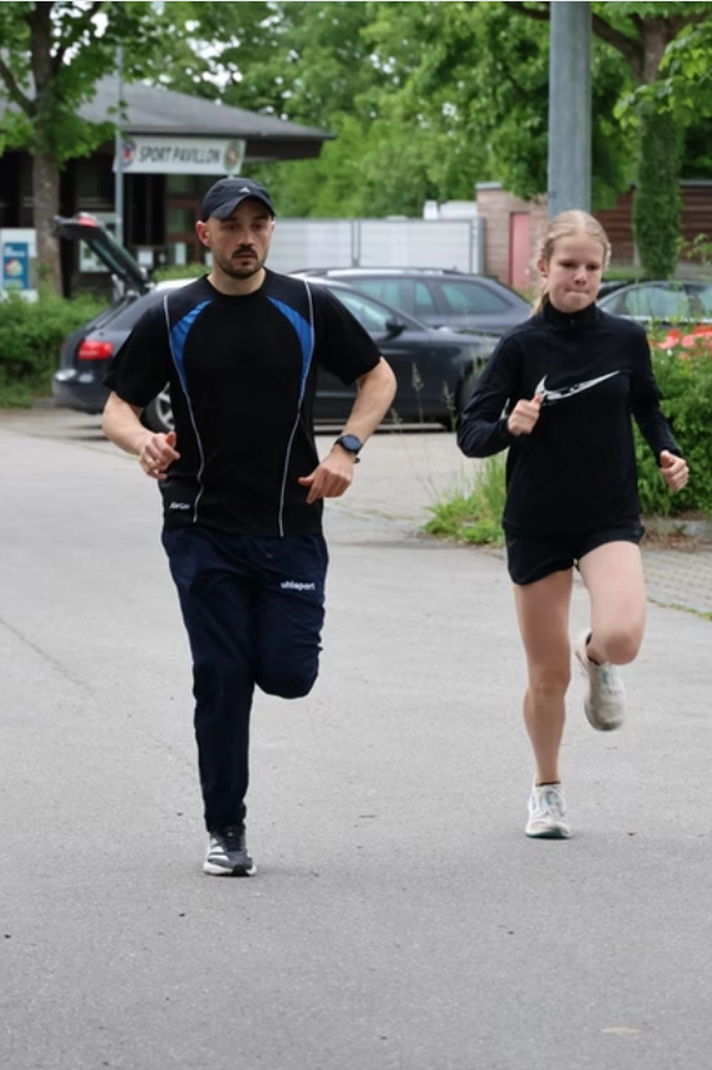 Der Laufcoach - Leistungscoaching und Analyse für Läufer - Ein Mann und eine Frau in Sportkleidung rennen gemeinsam auf einer asphaltierten Straße; im Hintergrund sind Autos und Bäume zu sehen.