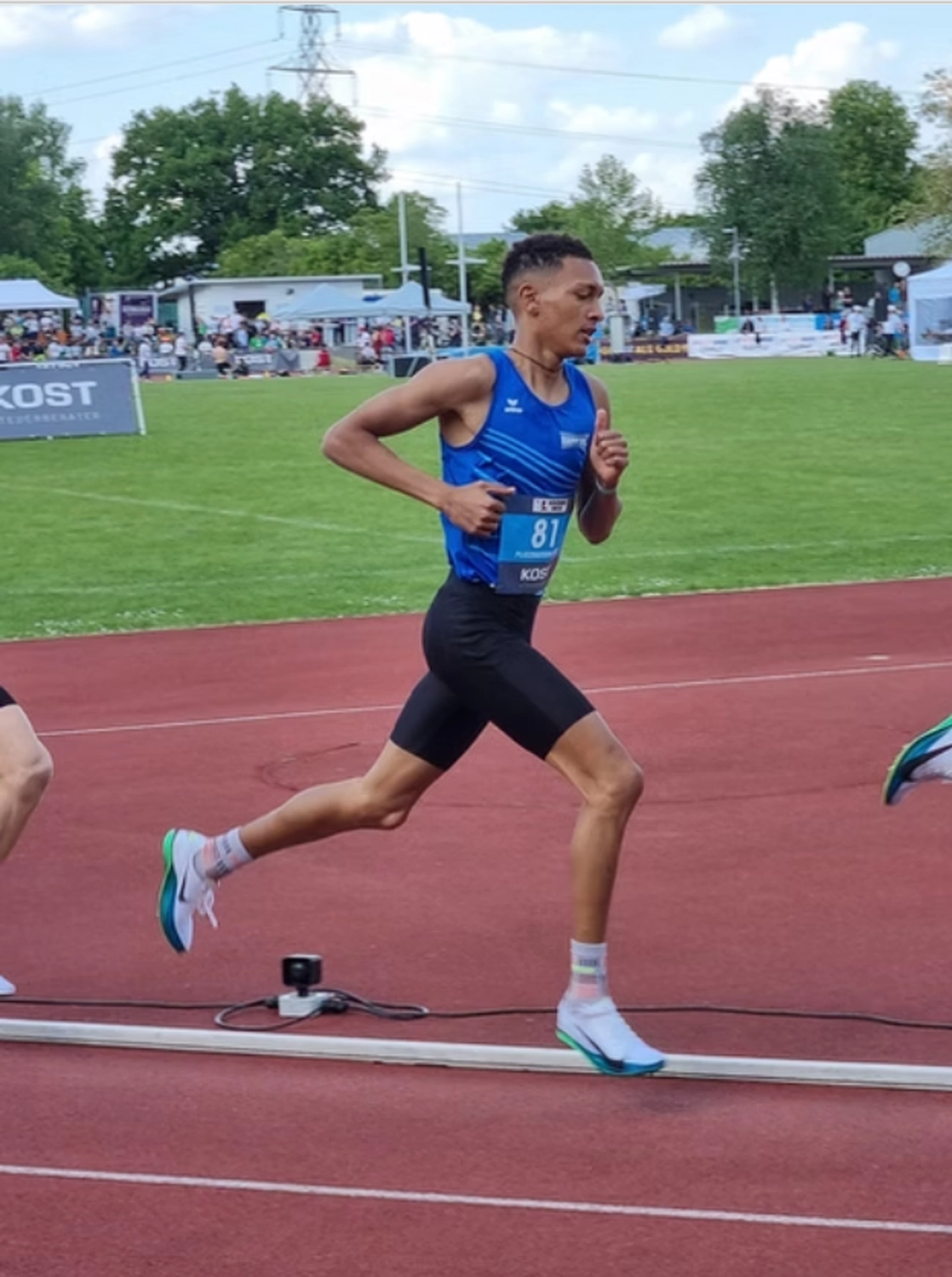 Der Laufcoach - Leistungscoaching und Analyse für Läufer - Ein athletischer junger Mann in einem blauen Tanktop und schwarzen Shorts nimmt an einem Leichtathletikwettkampf auf einer roten Freiluftlaufbahn teil.