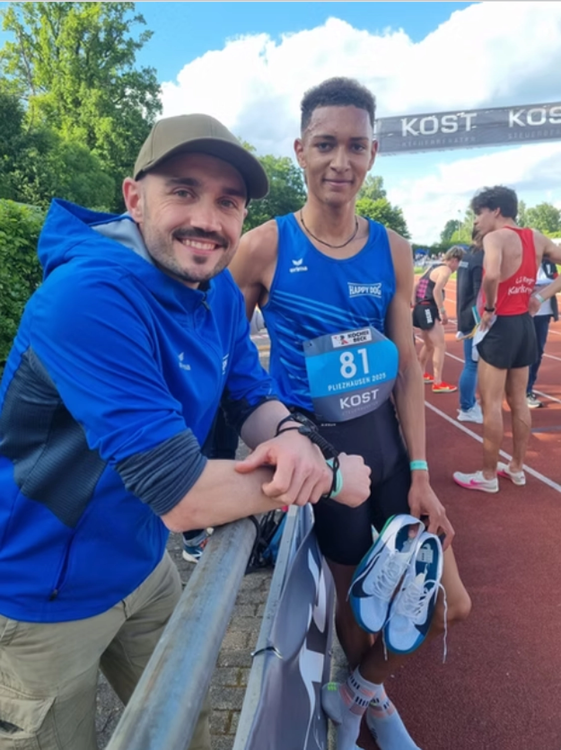 Der Laufcoach - Leistungscoaching und Analyse für Läufer - Ein lächelnder Mann in einem blauen Kapuzenpulli und einer olivgrünen Baseballkappe lehnt an einem Metallgeländer neben einem jungen Leichtathleten in einem blauen Tanktop, der seine Spikes hält.