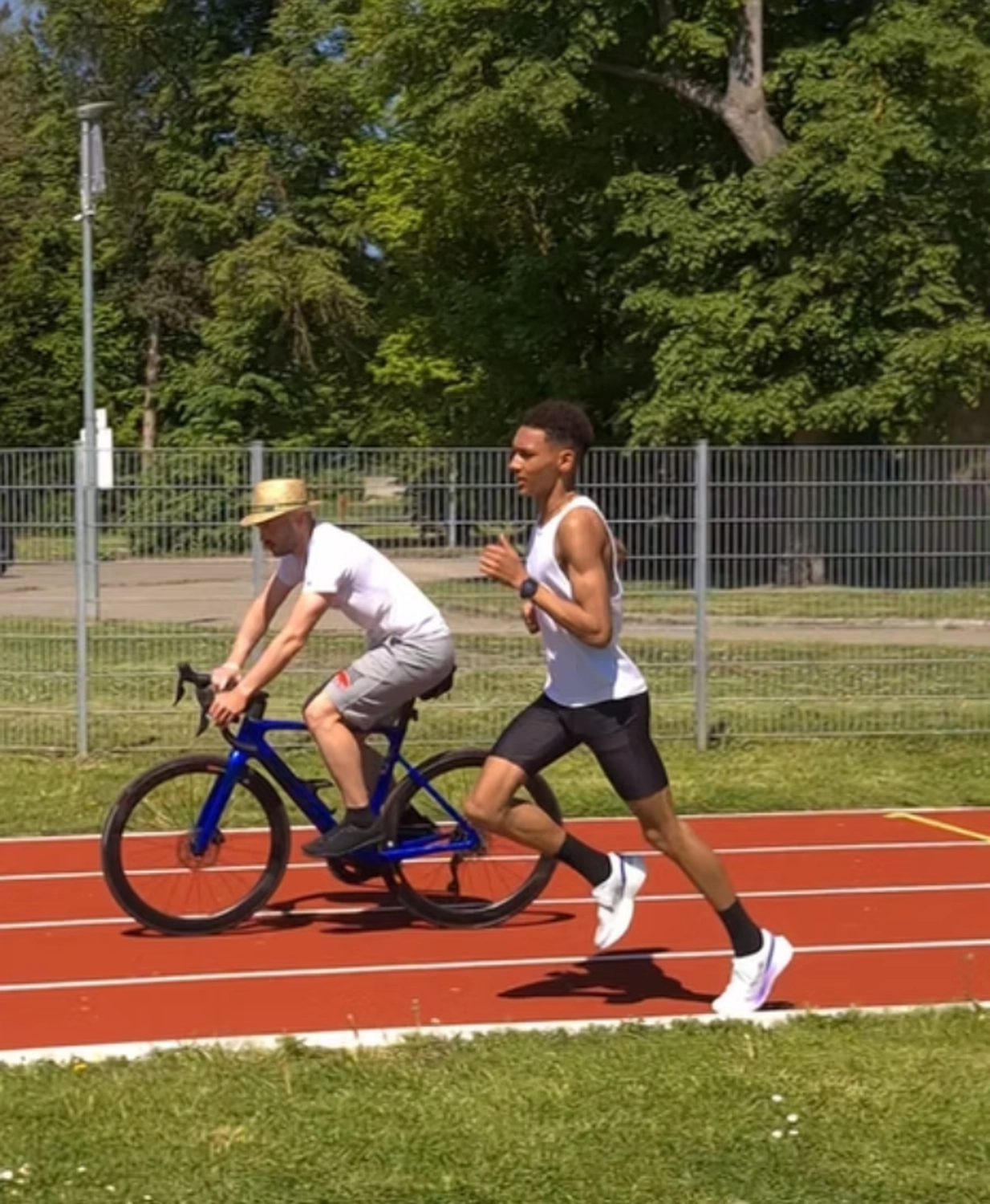 Der Laufcoach - Leistungscoaching und Analyse für Läufer - Ein junger Mann in Sportkleidung sprintet auf einer Freiluftlaufbahn, während ein anderer Mann mit Hut auf einem blauen Rennrad neben ihm herfährt.