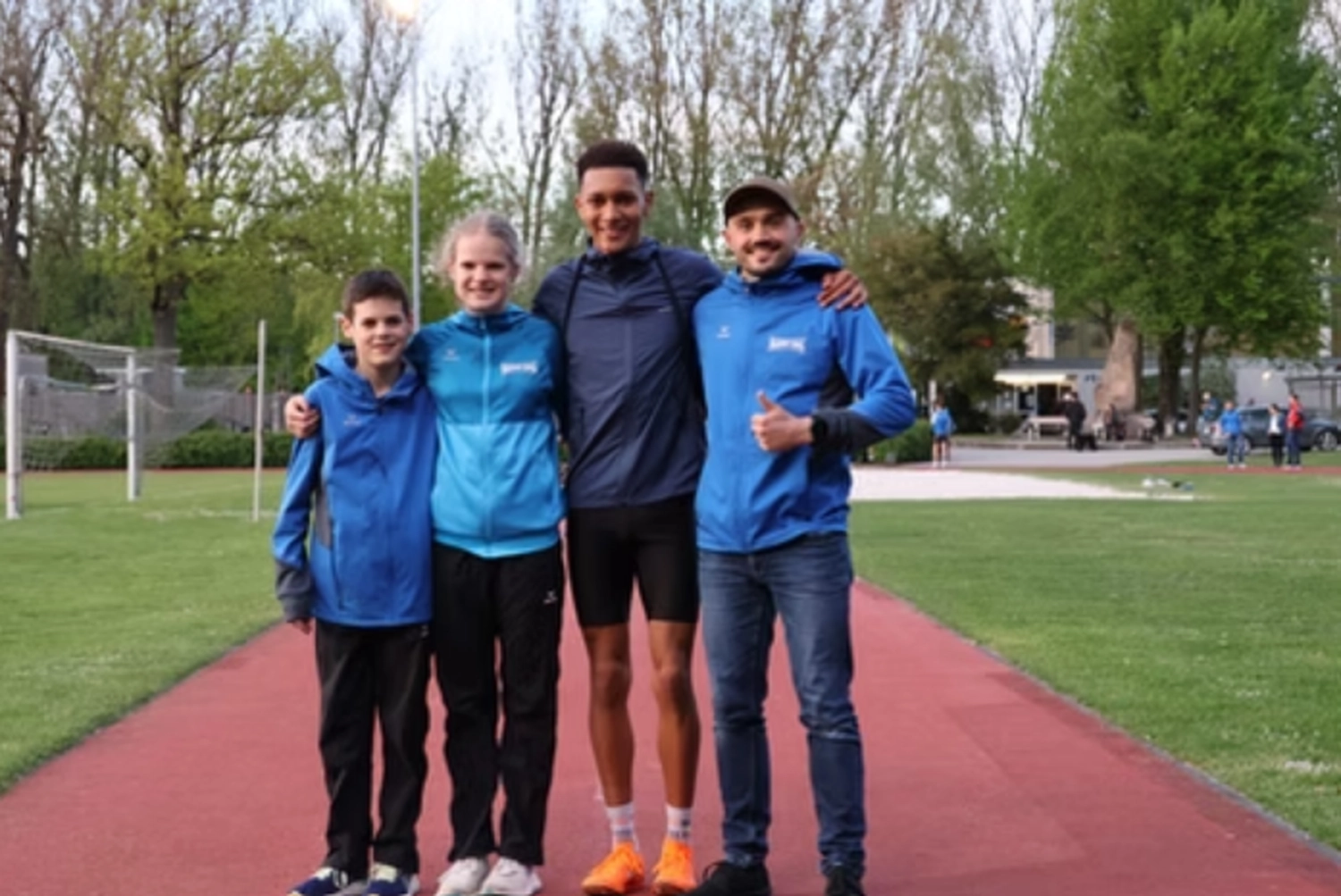 Der Laufcoach - Leistungscoaching und Analyse für Läufer - Vier Personen, darunter junge Sportler und zwei Erwachsene, stehen zusammen auf einer roten Laufbahn; im Hintergrund sind ein Fußballtor und grüne Bäume zu sehen.