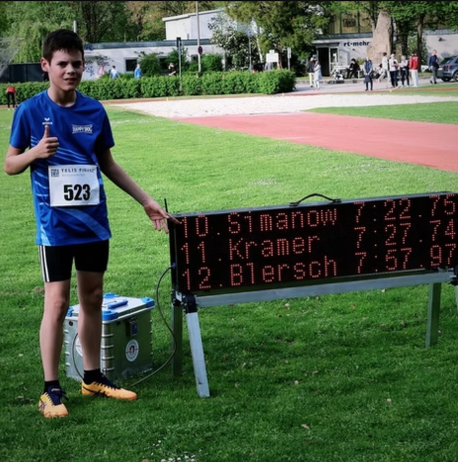 Der Laufcoach - Leistungscoaching und Analyse für Läufer - Ein junger männlicher Athlet in einem blauen Rennshirt und mit der Startnummer 523 steht auf einem Rasenfeld, zeigt mit erhobenem Daumen auf eine digitale Anzeigetafel, auf der die Rennergebnisse angezeigt werden.