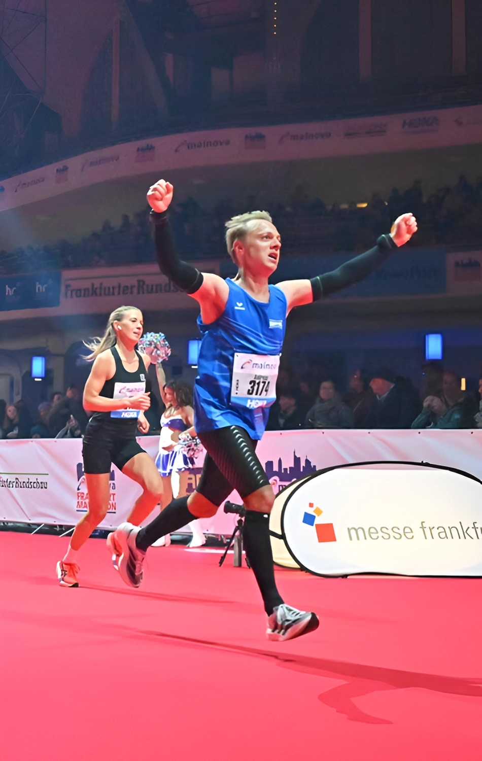 Der Laufcoach - Leistungscoaching und Analyse für Läufer - Ein männlicher Läufer in einem blauen Tanktop und schwarzer Kompressionskleidung jubelt mit erhobenen Armen, als er auf einem roten Teppich die Ziellinie überquert; im Hintergrund sind eine Läuferin und eine Menschenmenge zu sehen.