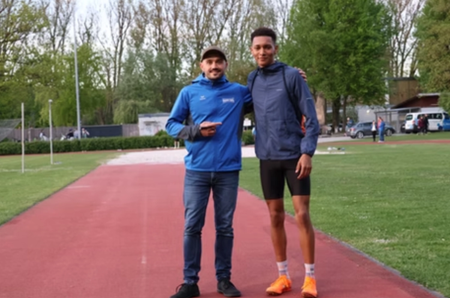 Der Laufcoach - Leistungscoaching und Analyse für Läufer - Zwei Männer posieren für ein Foto auf einer roten Freiluftlaufbahn; der eine trägt eine blaue Jacke und Jeans, der andere Sportkleidung und orangefarbene Laufschuhe.
