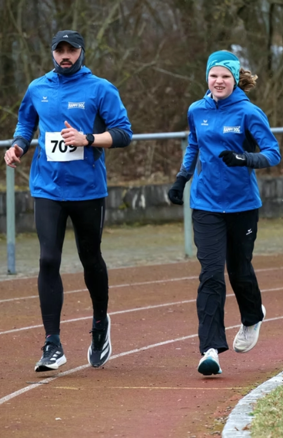 Der Laufcoach - Leistungscoaching und Analyse für Läufer - Ein Mann und ein Mädchen joggen Seite an Seite auf einer Freiluftlaufbahn; beide tragen die gleiche blaue Sportjacke und Sportbekleidung.
