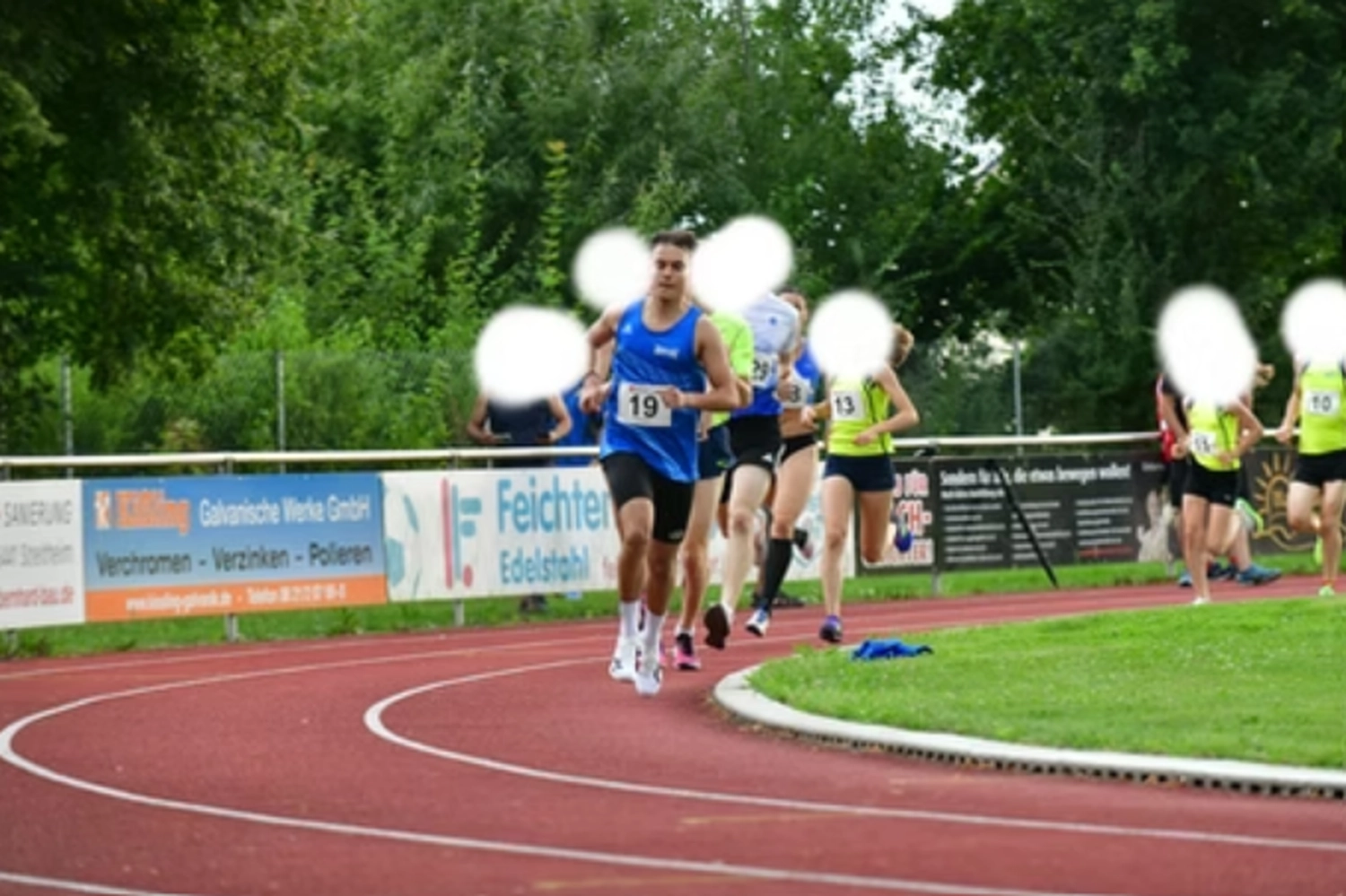 Der Laufcoach - Leistungscoaching und Analyse für Läufer - Ein Leichtathletikwettkampf im Freien mit mehreren Läufern in Sportkleidung, die auf einer roten Bahn, die von einem grünen Rasen und Werbebannern begrenzt wird, eine Kurve nehmen.
