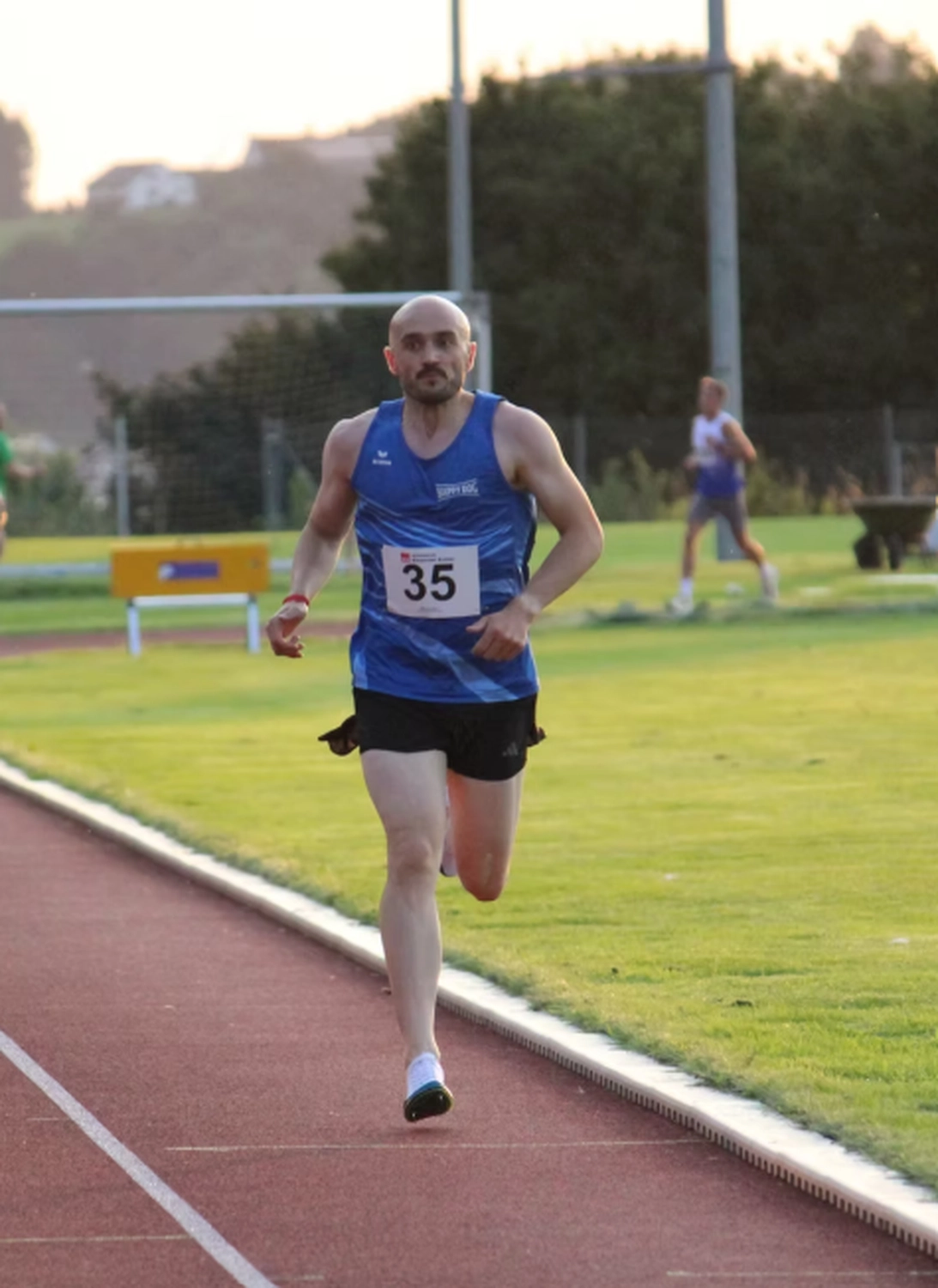 Der Laufcoach - Leistungscoaching und Analyse für Läufer - Ein kahlköpfiger männlicher Athlet in einem blauen Tanktop und schwarzen Shorts sprintet während eines Rennens eine rote Laufbahn entlang.