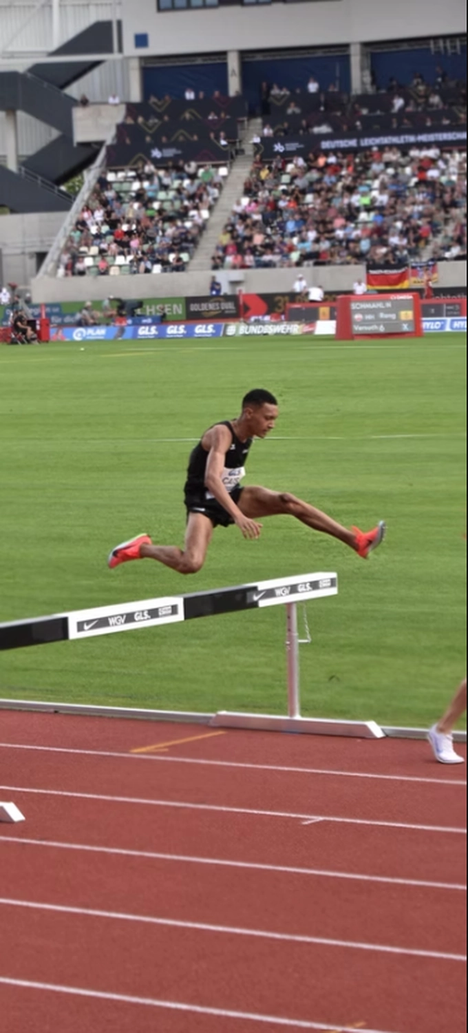Der Laufcoach - Leistungscoaching und Analyse für Läufer - Eine seitliche Aufnahme eines Leichtathleten in der Luft, wie er eine Hürde auf einer roten Laufbahn überwindet; im Hintergrund sind Stadionzuschauer und ein grünes Feld zu sehen.