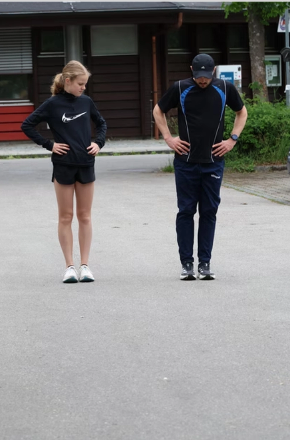 Der Laufcoach - Leistungscoaching und Analyse für Läufer - Ein junges Mädchen und ein Mann in Sportkleidung stehen auf einer gepflasterten Fläche, beide mit den Händen in den Hüften und den Blick auf den Boden gerichtet.