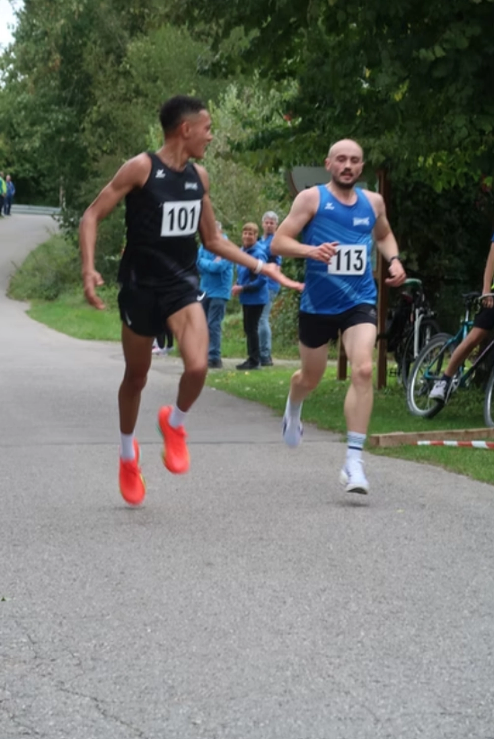 Der Laufcoach - Leistungscoaching und Analyse für Läufer - Zwei Läufer treten in einem Rennen auf einem gepflasterten Weg gegeneinander an, wobei ein Athlet in einem schwarzen Trikot (Nummer 101) vor einem anderen in einem blauen Trikot (Nummer 113) liegt.